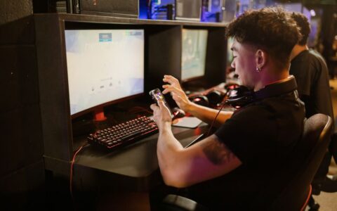 A young man is gaming using a smartphone and a PC setup with a mechanical keyboard indoors.