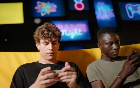 Two friends play mobile games indoors, focusing intently on their smartphones.