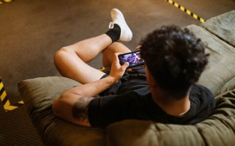 A man relaxing on a sofa playing a video game on his smartphone.