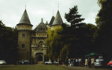 A beautiful stone castle with vintage cars and people gathered outdoors on a cloudy day.