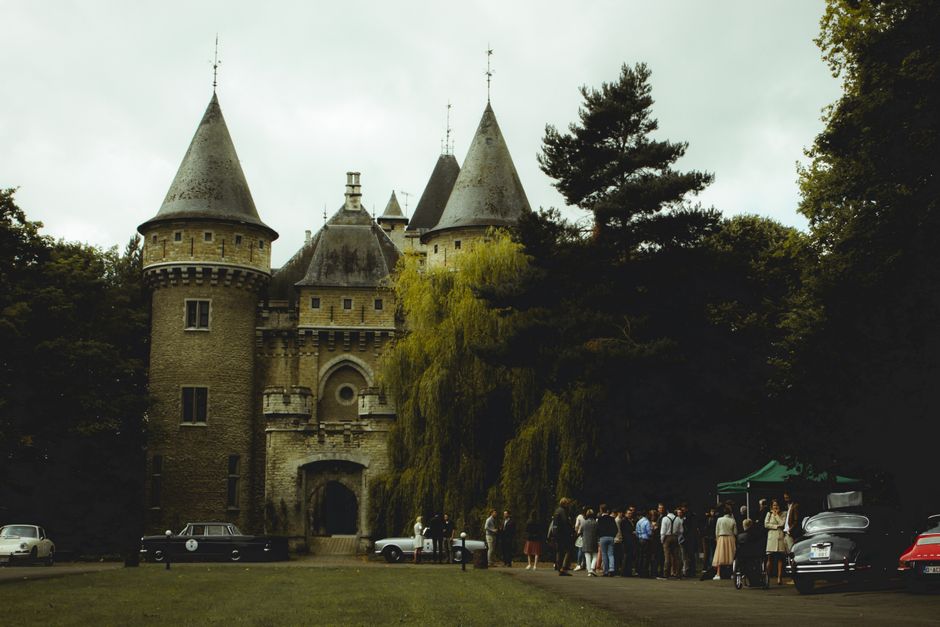 A beautiful stone castle with vintage cars and people gathered outdoors on a cloudy day.